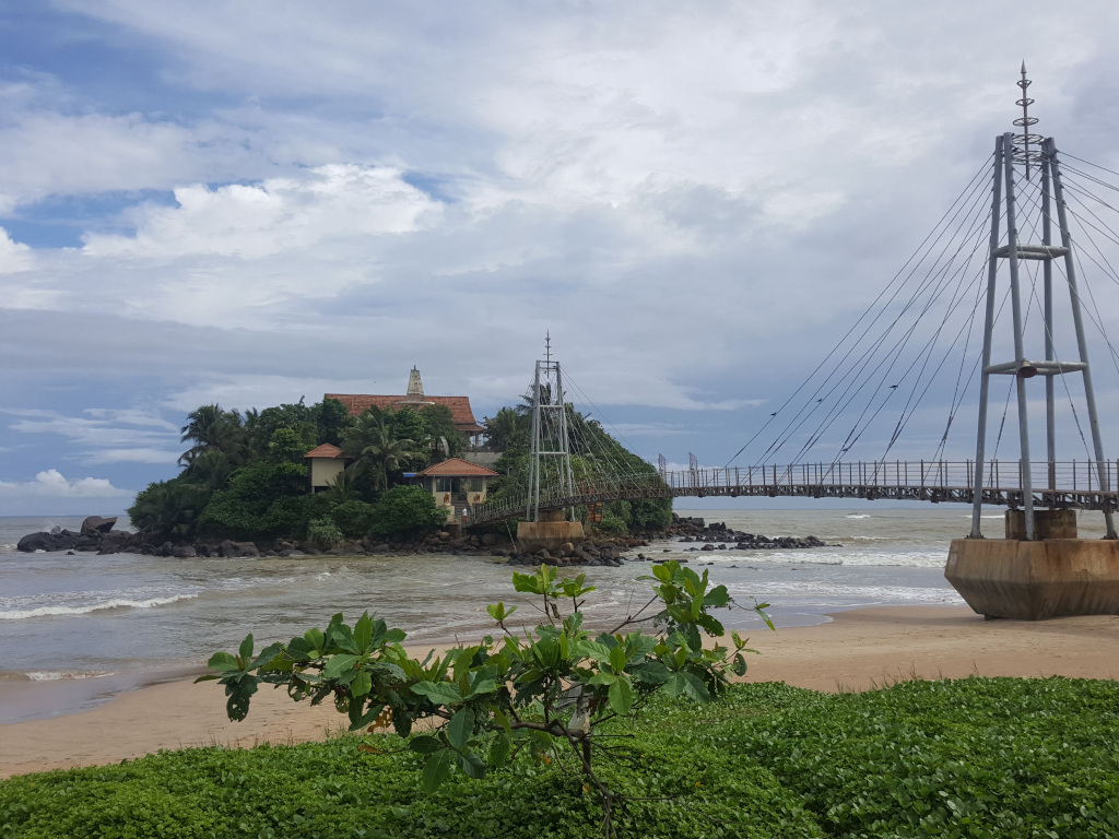 Small, verdant island just off a sandy beach, connected to the mainland by a modern, slender suspension bridge. The island features a cluster of buildings with reddish-brown tile roofs, suggesting a temple complex or religious retreat nestled amongst lush tropical vegetation, including palm trees. The bridge is metallic gray, with thin cables supporting a narrow walkway. The bridge's pylons are anchored to concrete bases that partially emerge from the sand. There are no people visible. A few birds are perched on the cables of the bridge.