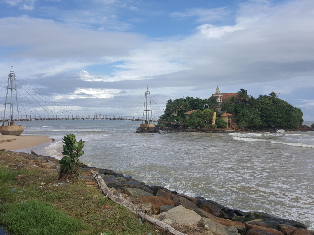 Small, verdant island connected to the mainland by a modern, slender suspension bridge. The island is densely covered with tropical vegetation, punctuated by a light-brown, multi-level building, appearing to be a temple or shrine, with a visible spire. The bridge is a pale grey-blue, its cables elegantly arching against the sky. The water surrounding the island is a muted grey-green, with small waves gently lapping the shore. On the mainland, a rocky shoreline, strewn with debris and sparse vegetation, including a small, bright green succulent plant, runs alongside the beach. No people are visible.