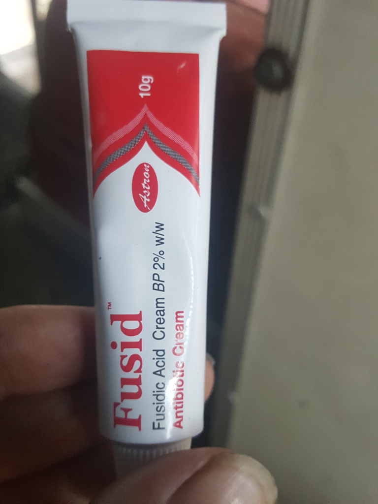 Tube of Fusid antibiotic cream, held gently between the thumb and fingers of a light-skinned hand. The tube is predominantly white, with a vibrant red band at the top displaying the brand name Astron in a stylised script font and 10g indicating the quantity. The tube is slightly angled toward the viewer.