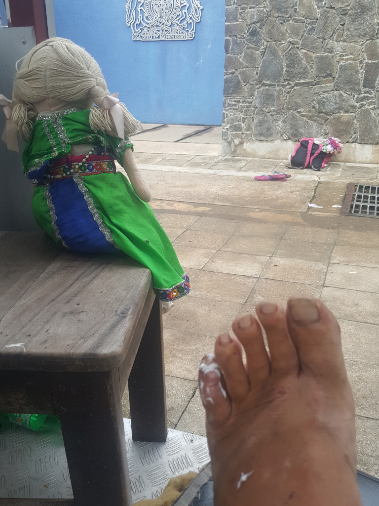 Slightly low-angle, close-up shot featuring a weathered, bare foot in the foreground, prominently displaying a large amount of white ointment or cream on the big toe. The foot appears tanned and calloused, indicating manual labour or outdoor exposure. In the mid-ground, slightly out of focus, sits a handmade doll dressed in a vibrant green and blue garment, reminiscent of traditional Indian or South Asian attire. The doll is positioned on a worn, dark brown wooden stool, its back to the viewer. Its light-coloured yarn hair is styled in pigtails with pale pink bows. A pink backpack, seemingly casually discarded, lies on the ground in the background to the right of the doll. A pair of sandals rest nearby. The setting appears to be an outdoor area, possibly a courtyard or passageway. A wall constructed of rough, grey-brown stones forms the backdrop to the right. To the left, a faded blue wall is visible, bearing a worn, royal crest or insignia which suggests a place of authority or historical significance. This wall also suggests some official or government type building. The ground is paved with light-grey concrete tiles. A drainage grate is visible in the lower right corner. The overall lighting is soft and diffuse, likely daytime natural light, casting no harsh shadows.