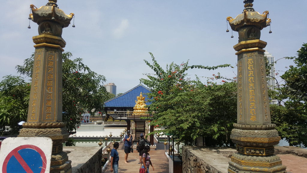 Pathway leading towards a golden Buddha statue nestled within a blue-roofed structure. This structure appears to be a temple or shrine. Flanking the path are two imposing, ornate gray stone pillars, richly detailed with gold lettering and carvings that resemble Buddhist scriptures. The pillars are capped with elaborate, tiered roofs adorned with small bells. Several people are visible on the path, primarily positioned near the base of the golden Buddha. They are casually dressed in modern clothing; one man wears a teal shirt, another a dark gray shirt. A woman in a dark floral-printed shirt and another woman in a light-coloured shirt and dark pants are standing near the centre. A young child is also present. The people appear to be observing the statue, seemingly unhurried and contemplative. The scene is bathed in bright, natural sunlight, suggesting a daytime setting with clear skies. The light casts soft shadows, enhancing the textural details of the stone and the metallic gleam of the gold Buddha. Lush green foliage, comprised of trees and bushes with hints of reddish flowers, frames the pathway and partially obscures the background. The background reveals a modern cityscape, featuring several tall buildings under a pale blue sky, offering a juxtaposition of traditional architecture and modern urban life. The blue roof of the shrine contrasts sharply with the gold Buddha, and the dark gray stone of the pillars adds depth and solidity to the composition.