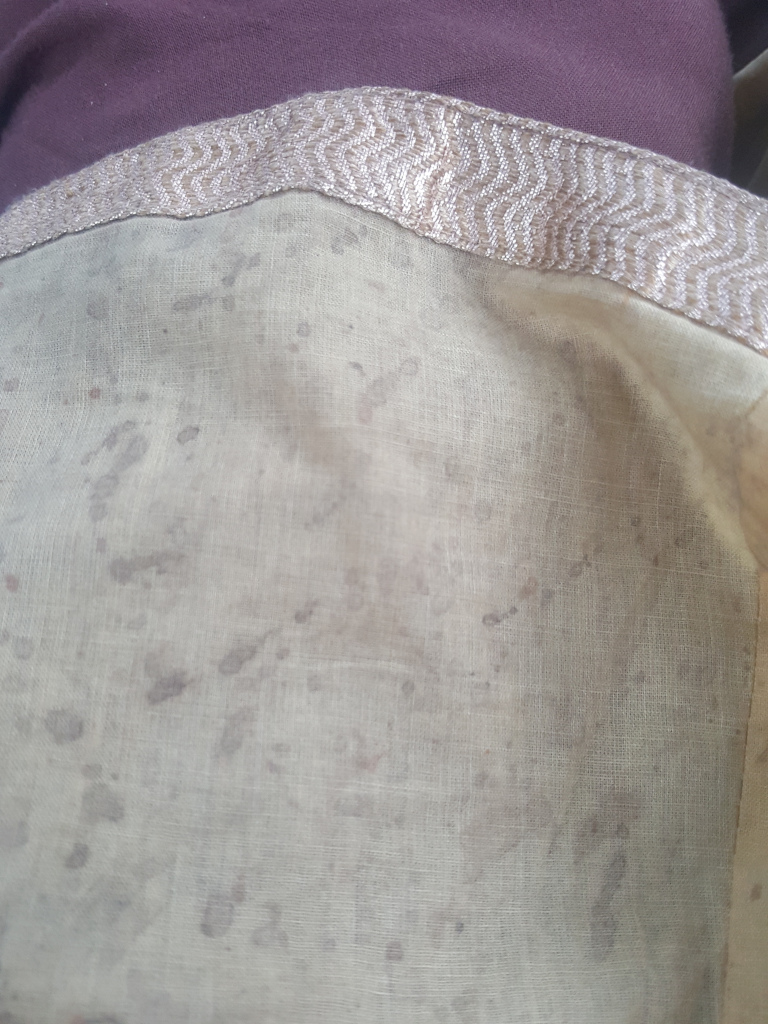 Textured linen, which appears soft and slightly sheer. The stains are scattered across the surface, varying in size and intensity, giving a mottled effect. A band of decorative trim is visible at the top edge of the linen. This trim is a light beige/off-white with a subtle, repeating chevron or zigzag pattern woven into it, showing a slight shimmer suggesting metallic thread. The trim is positioned horizontally, marking a clear separation between the linen and an unseen background. Part of a dark purplish-brown garment is visible just above the trim. he background is largely obscured, showing only a sliver of a dark brownish-purple fabric, suggesting the linen is part of a larger garment or drape. The lighting is soft and diffuse, with no harsh shadows, suggesting either indoor lighting or indirect sunlight. The colour palette is muted and earthy, dominated by the light beige of the linen, the darker brown of the stains, and the dark purplish-brown of the background fabric. The silver-toned threads in the trim provide a subtle contrast.