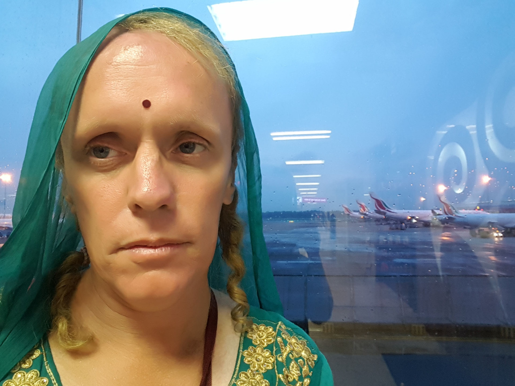 Leonie with light blonde hair, partially obscured by a teal-green silk dupatta (scarf) draped over her head and shoulders. The dupatta is loosely gathered, allowing strands of her hair to peek out from the sides, which are styled in loose braids. She wears a teal-green kurta (tunic) embroidered with gold floral motifs around the neckline and shoulders. A small, dark red bindi (forehead dot) adorns her forehead between her eyebrows. Her expression is serious, almost pensive, with slightly downturned eyes and lips, suggesting a thoughtful or perhaps slightly melancholic mood. She appears to be sitting or standing still, gazing slightly off to her left, toward the background. The background is a large, slightly rain-streaked window offering a view of an airport tarmac at dusk or dawn. Multiple large passenger jets, predominantly white with red and blue markings (possibly Sri Lankan Airlines), are visible parked on the tarmac. The lighting is soft and diffuse, characteristic of early morning or late evening light, creating a muted, slightly bluish overall cast. The airport lights are visible in the background as are faint lights from the terminal itself. The colors are predominantly muted: teal-green of the dupatta, the gold embroidery on the kurta, the pale skin tone of Leonie, and the greyish-blue tones of the sky and wet window. The overall palette is subdued and somewhat melancholic.