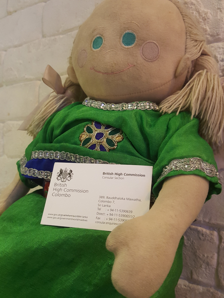 Rag doll, appearing handmade, with a light beige, almost off-white face, rosy cheeks subtly painted on, and bright turquoise eyes. Its blonde yarn hair is styled in two loose pigtails secured with simple bows. The doll is dressed in a vibrant emerald green, ankle-length garment, adorned with gold and purple embroidery around the neckline and a decorative floral motif at the chest. The fabric appears soft and slightly textured. The doll's arms are slightly bent, and it gently holds a business card in its hands. The business card, crisp and white, prominently displays the British High Commission, Colombo logo and contact details. The doll sits slightly angled, presenting the card slightly towards the viewer.