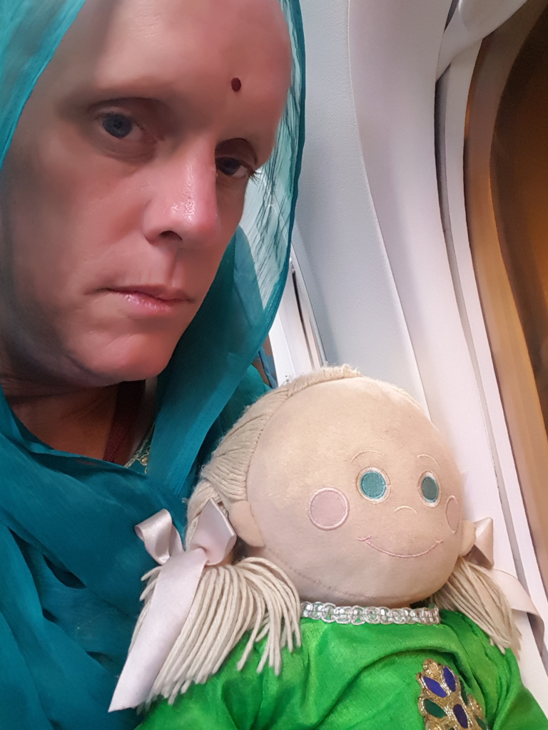 Close-up shot of a Leonie, her face slightly angled to the left, partially obscured by a vibrant teal, sheer dupatta (a type of scarf) draped over her head and shoulders. The woman appears to be middle-aged, with fair skin, visible pores, and a small, dark red bindi (a decorative mark) centred on her forehead. Her expression is serious, almost melancholic, with slightly downturned lips and a hint of sadness in her eyes. Her hair is not visible beneath the dupatta. In her arms, she gently holds a soft, rag doll. This doll is light beige or cream-coloured with rosy cheeks, embroidered light blue eyes, and a subtly stitched-on smile. It has long, stringy, light blonde hair styled in pigtails, each secured with a pale pink ribbon bow. The doll wears a shimmering, emerald green dress with gold detailing near the neckline.