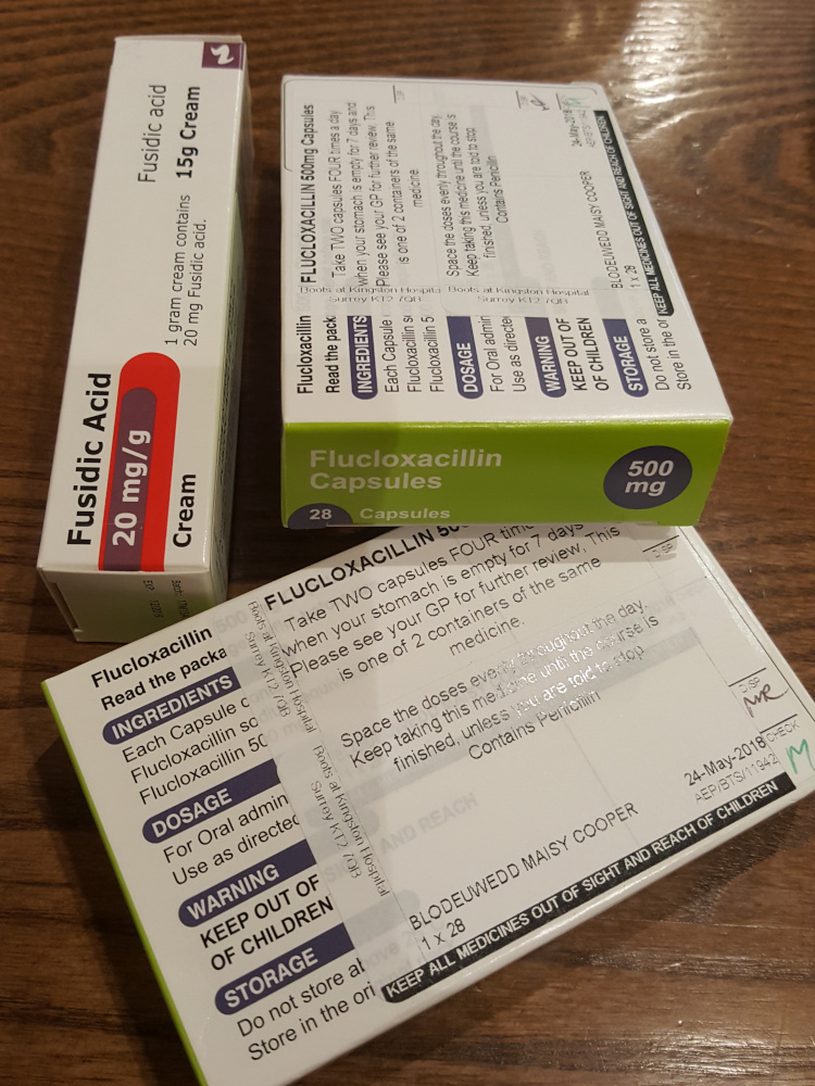 Three pharmaceutical packages resting on a dark brown surface, likely a table or counter top. A lime-green and white box of Flucloxacillin 500mg capsules (28 capsules). The box is partially angled, revealing the side and top panels with detailed text about dosage, ingredients, warnings, and storage. The text is clearly printed in black lettering on a contrasting background. The box shows wear and creasing. A slightly smaller, light-blue and white box of Flucloxacillin 500mg capsules (28 capsules). It's oriented horizontally, showing its long side and the top, revealing similar information to the green box, with smaller text size. The packaging also bears signs of handling and slight damage. A white and light-blue tube of Fusidic Acid 20mg/g cream (15g). This tube is slightly tilted. The label clearly indicates the product's name, concentration, and content amount. The text is sharp and well-defined. All boxes appear to be used and have some level of wear, implying they were previously opened.