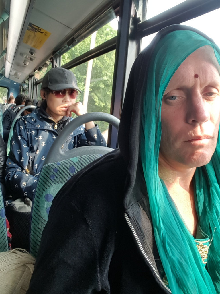 Two women seated on a public bus. Foreground Woman: Dominates the foreground, wearing a dark navy or black hooded sweatshirt over a teal, draped dupatta (a long scarf worn by South Asian women). The dupatta's fabric has a slightly rippled texture and is loosely wrapped around her neck and shoulders. Her face is visible, showing pale skin with some freckling or sunspots, and she has a somber, almost weary expression. Her eyes are slightly downturned, and her lips are pressed together in a neutral or slightly unhappy line. There's a small, dark red bindi (forehead dot) visible on her forehead. Her hair is concealed entirely under the hooded sweatshirt and dupatta. Background Woman: Seated slightly behind and to the left of the foreground woman, this woman is wearing a navy blue jacket with small anchor motifs and a dark-colored baseball cap. She has sunglasses perched on her nose, and she's leaning slightly to her left with her hand resting on her chin, suggesting a contemplative posture. Her expression is somewhat reserved and distant.