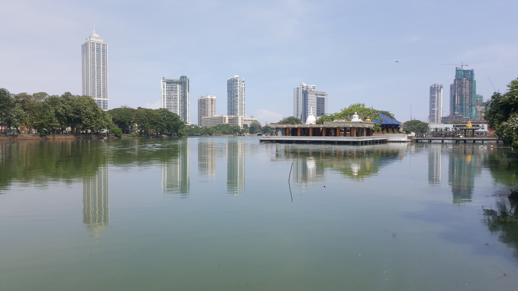 Calm, greenish-grey lake reflecting a cityscape and a modern, light-brown structure. The structure, situated centrally in the lake's middle ground, appears to be a pavilion or temple with a flat roof and distinct pillars.  It has a white, slightly elevated central section. Surrounding the lake are lush, dark-green trees forming a natural border along the shoreline. In the background, several modern skyscrapers of varying heights rise against a pale, almost cloudless sky. One particularly tall, slender skyscraper dominates the left side. Another, more blocky building is under construction with a visible crane. These buildings are rendered with sharp lines and distinct architectural details. There are no people visible in the image.