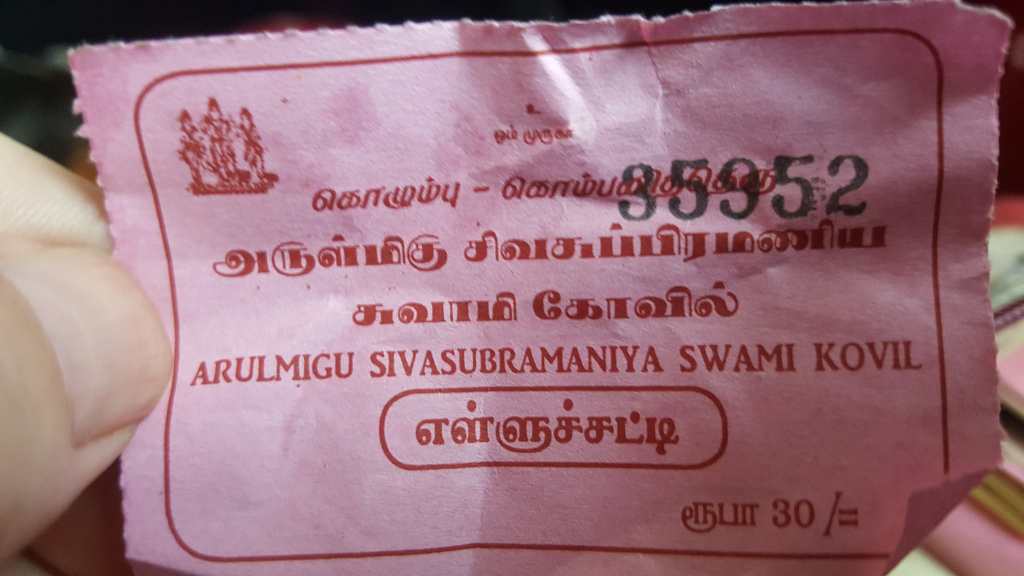 Crumpled, pinkish-tan ticket, held gently between the thumb and forefinger of a light-skinned hand. The ticket itself is rectangular with slightly irregular edges, suggesting it's been torn from a larger roll. The ticket's text is predominantly dark red-brown, printed in both Tamil and English. A small, simple woodcut-style image of what appears to be a temple or religious scene sits near the top left. The number 35952 is prominently displayed, appearing almost as a serial number.