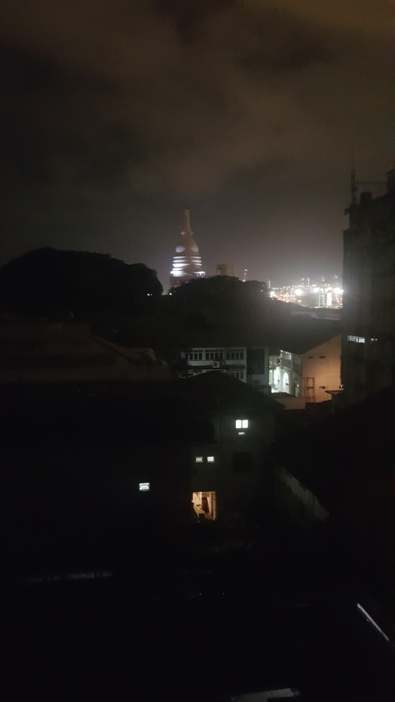 Distant, brightly lit stupa, a dome-shaped Buddhist monument, standing tall and slender against the night sky.  It's centrally positioned, but slightly off-center, creating a subtle visual imbalance. In the foreground, several dark, silhouetted buildings are visible, their outlines suggesting residential structures with dimly lit windows.  The buildings are rendered almost entirely in shadow, with only a few internal lights breaking through the darkness. A portion of a larger, possibly multi-story structure, is visible on the right edge of the frame, also partially obscured by darkness.