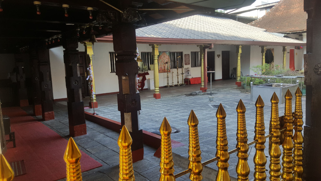 Courtyard scene within what appears to be a Hindu temple or similar religious structure. The scene is set outdoors, in a relatively enclosed courtyard within a temple complex. The background shows a section of a larger building, possibly part of the temple or an attached structure. The lighting is soft and diffused, likely natural daylight, suggesting an overcast day or shaded environment. The colour palette is fairly muted; the dominant colours are the soft grey of the stone paving, the dark brown of the wooden pillars, the creamy white of the building walls, and the striking gold accents of the fence and some of the temple's adornments. The red carpet adds a small pop of deeper colour.