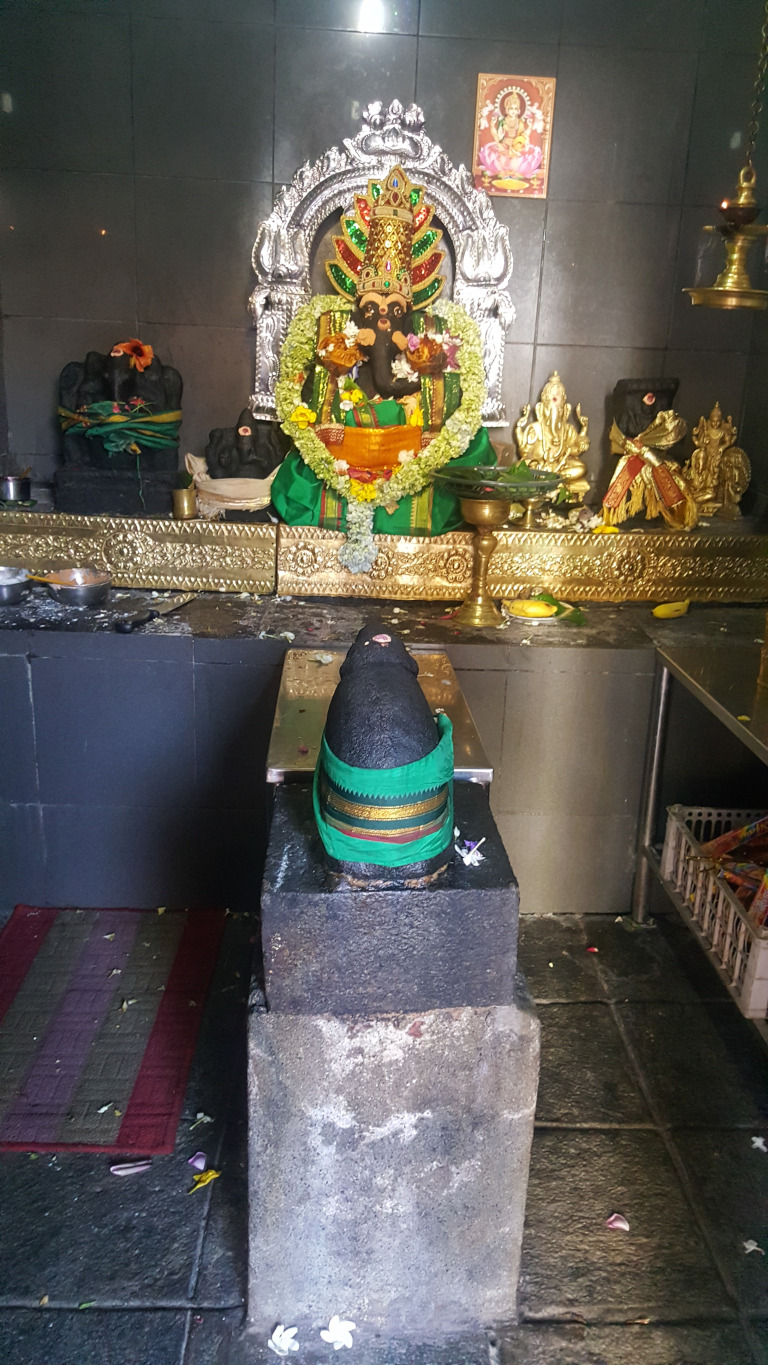 Dark gray, almost black, roughly hewn stone lingam (phallic symbol) on a square, weathered gray stone pedestal. The lingam is adorned with a vibrant green and gold sash, neatly wrapped around its midsection. It sits slightly elevated above the floor, directly in the foreground. Above it, the main deity is a richly decorated statue of Ganesha, seated on a silver throne with ornate carvings. Ganesha's attire is lavish; a bright green and orange robe with abundant floral garlands. The deity features a dark, almost black complexion and intricate headdress with sparkling gold and green details. Flanking Ganesha are smaller statues, appearing to be additional deities or aspects of the divine, also adorned with gold and vibrant colours. At the far left and right of the main shrine, are dark gray stone representations of other Hindu deities, likely Ganesha (with elephant heads), draped in green cloths.