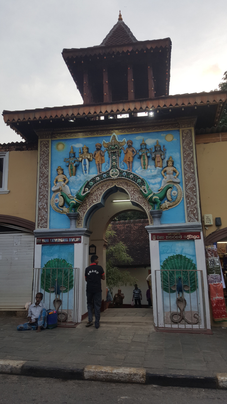 Vibrant, intricately decorated entranceway to what appears to be a Sri Lankan temple or shrine. Dominating the scene is a large, arched gateway, painted in a predominantly blue hue, showcasing a lively depiction of Hindu deities in a celestial scene. The deities are rendered in rich, jewel-toned colours, with gold accents highlighting their clothing and ornamentation. Flanking the arch are painted murals of peacocks perched atop cobras, symbols of both beauty and protection. Directly beneath the archway, a man in a dark, short-sleeved shirt with Chinese characters stands facing away from the camera, his posture relaxed yet attentive. To the lower left, an older man sits on the ground, dressed in simple, light-coloured clothing, his gaze slightly lowered. He appears to be resting or waiting. In the background, near the archway's inner passage, several other figures are visible, but their details are obscured by distance and shadow. These figures suggest other visitors or possibly temple attendants.