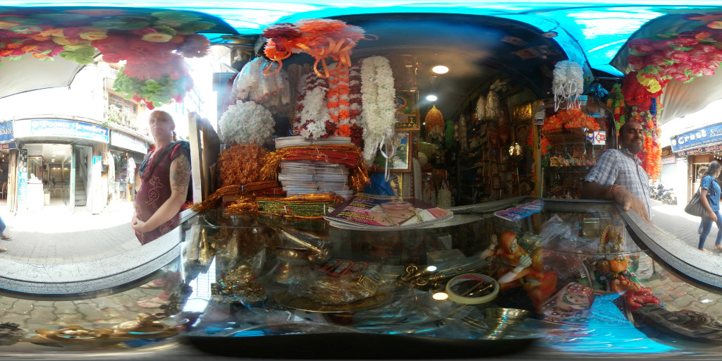 Leonie with fair skin and tattoos on her left arm stands slightly off-centre left, facing the viewer. She wears a maroon, patterned long-sleeved top. Her expression is neutral, almost pensive. Her posture suggests a quiet observation of the scene. A man with brown skin and glasses stands behind a glass counter on the right, seemingly attending to his shop. He wears a plaid, light blue and white shirt. His expression is relaxed and welcoming. The central focus is a glass display case overflowing with vibrant garlands, mostly artificial flowers in bright orange, red, white and yellow hues. Stacks of what seem to be colourful fabrics or papers are piled behind the garlands. Smaller religious figurines and metal objects are also displayed within the case. A roll of tape is visible among the items.