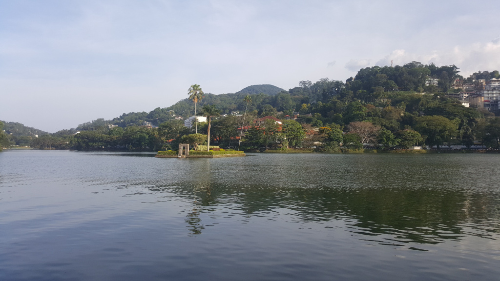 Serene lake, its dark, still water reflecting the surrounding landscape. A small, verdant island sits prominently in the mid-ground. This island features lush tropical vegetation, including several palm trees, and a small, aged-looking stone structure, possibly an arch or a small pavilion, near its centre. The structure appears to be made of light-grey or tan stone, weathered by time and exposure to the elements. There are no people visible in the image. The scene is set in a tropical or subtropical climate. The background is dominated by a gently sloping hill densely covered with lush green vegetation.  Houses and buildings are interspersed within this greenery, appearing mostly as residential structures, scattered among the trees. The hills rise smoothly to meet the sky. The sky is a soft, light blue, with just a hint of cloud cover, suggesting a calm, possibly slightly overcast day. The lighting is soft and diffused, indicative of either early morning or late afternoon light. There are no harsh shadows. The overall colour palette is muted yet vibrant: dark greens of the foliage, darker greys and browns of the buildings and stone structure, the dark grey-blue of the calm lake, and the pale, washed-out blue of the sky.