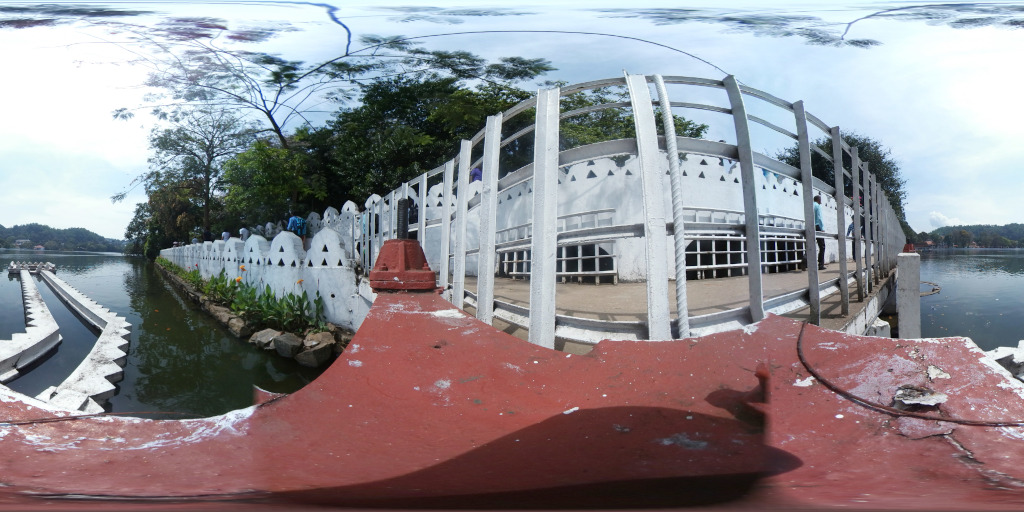 White, balustraded walkway that extends from the lower-middle foreground into the middle distance, curving gently to the right. The walkway borders a serene body of water, seemingly a lake or pond, that stretches across the lower-middle and lower-background. The walkway's white balustrade is punctuated by decorative arched sections in a scalloped pattern. A reddish-brown, weathered metal structure forms the foreground, appearing as a part of the walkway's supporting structure. It shows signs of wear and tear, with patches of peeling paint and visible metal. The walkway itself has several people walking on it; they're small figures, their actions and details indistinguishable. On the opposite side of the walkway, the ground meets the water; a white, segmented structure runs parallel to the walkway in the water at the far left edge.