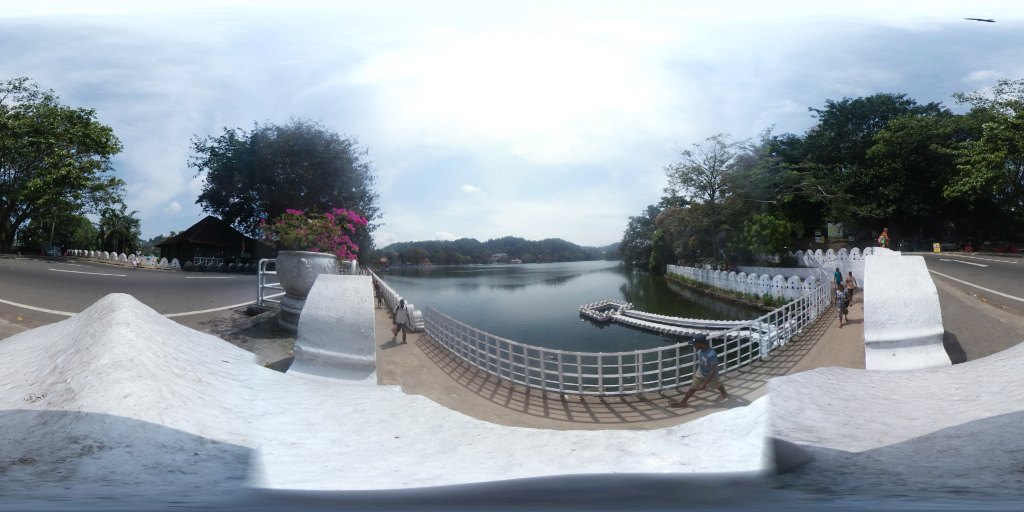 Picturesque bridge spanning a serene lake. The bridge is characterised by a high, white, textured wall along its sides, punctuated by regularly spaced, slightly rounded, white pillars. The wall's texture is rough and slightly uneven, not perfectly smooth. A simple metal railing runs along the bridge's edge, providing a boundary between the walkway and the water. Several individuals are scattered along the bridge's walkway. Most are children, walking in small groups or individually, seeming casually dressed in light-coloured clothing. One adult is visible, dressed in what appears to be a light-coloured shirt and pants, near the centre of the scene. These figures are small in the overall perspective, giving a sense of scale to the scene. Two boats or floating platforms are visible in the lake, near the bridge. A large, ornate stone planter containing vibrant pink flowers sits on the bridge, near the left side of the frame.