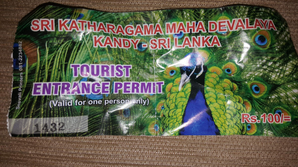 Crumpled, slightly dirty tourist entrance permit. The permit is rectangular, with its edges showing signs of wear and tear, particularly at the top left and bottom corners where it appears creased and frayed. The overall texture suggests it's made of thin, possibly laminated, paper or plastic. The permit itself is the dominant object, occupying almost the entire frame. The text on the permit is predominantly in a bold, sans-serif font, appearing in shades of purple and red against a background image of a vibrant peacock. The peacock's tail feathers are spread out, showcasing their intricate pattern of blues, greens, and golds.