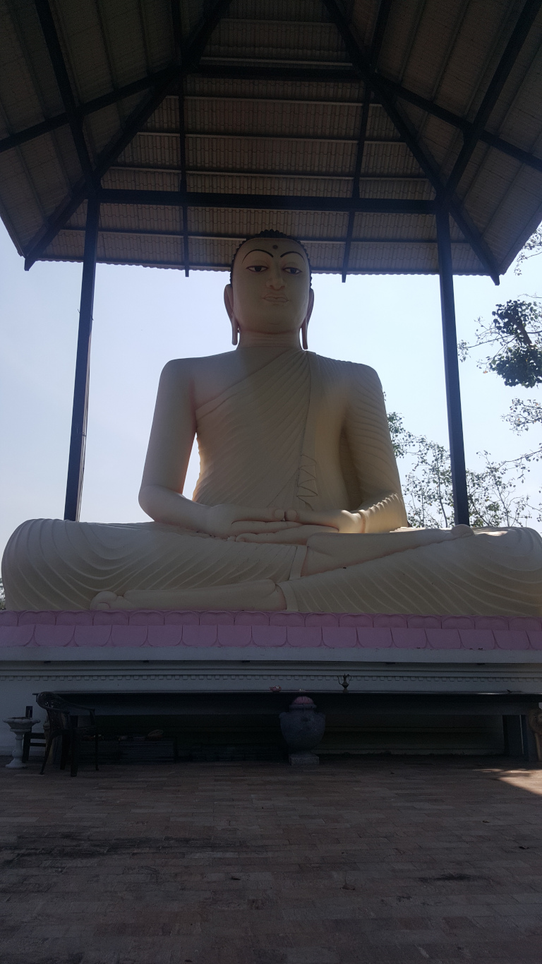 Large, serene statue of Buddha, depicted in a seated meditation posture (dhyanasana). The Buddha is light beige or cream-coloured, with smooth, subtly textured robes draped elegantly. The facial features are calm and peaceful, with a slight, gentle smile hinted at the corners of the lips. The statue's size is imposing, dwarfing the surrounding structure. The Buddha's hands rest gently in his lap. The statue sits on a low, pink platform.
