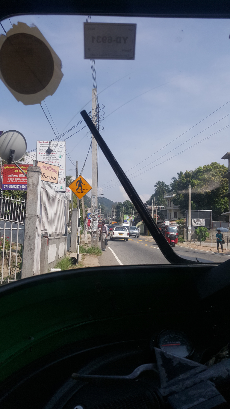 Inside of a three-wheeled vehicle, likely a tuk-tuk, looking out through the wind shield. The wind shield is partially obscured by a noticeable circular sticker, torn at the edges and showing a faded, tan under layer. A smaller, rectangular sticker reading Y033-DY is neatly affixed to the upper right. The vehicle's interior is dark and mostly obscured. A portion of the speedometer is visible at the bottom, hinting at a muted red needle.