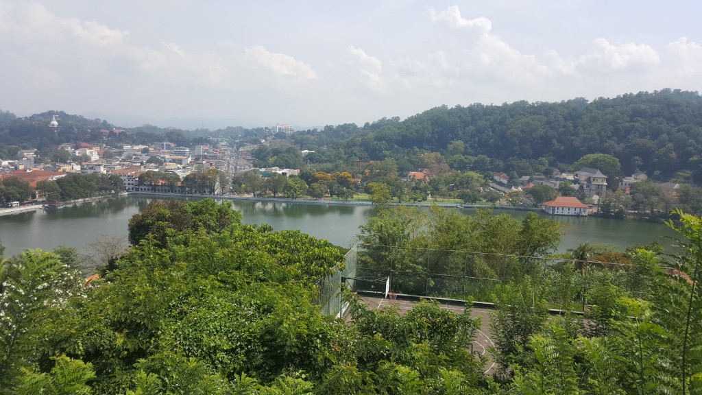 Panoramic view of Kandy Lake, Sri Lanka, nestled within a vibrant green landscape. The lake itself is a calm, dark teal, reflecting the surrounding sky and foliage. A portion of a basketball court is visible in the foreground, partially obscured by lush vegetation. The city of Kandy sprawls across the background, showcasing a mix of low-rise buildings in muted earth tones, punctuated by occasional white structures.  Hills heavily forested with deep green trees rise gently beyond the lake and the city, creating a layered backdrop. A white structure, possibly a stupa or temple, is faintly visible on a distant hill to the left. There are no people visible in the image.