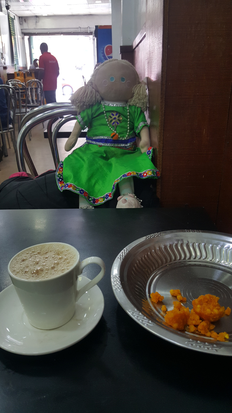 Dominating the foreground are a white cup and saucer holding a light brown, frothy beverage (likely tea or coffee), positioned slightly left of centre. Beside it, a shallow, silvery-grey metal plate with ornate edging holds remnants of a vibrant orange, crumbly confection—possibly a type of Indian sweet. A rag doll, centrally positioned but slightly off-center, is seated on a stainless steel chair. The doll is striking, with a light tan face, light brown yarn hair styled in pigtails, and a bright green dress adorned with colourful embroidery and beading along the neckline and hem. It wears what appears to be silver jewellery. The background is blurry but reveals the interior of what seems to be a bustling South Asian-style cafe or restaurant. There are metallic chairs, tables, and other patrons (partially visible) in the distance. A man in a red shirt is visible near the back, suggesting a staff member. A doorway leads to the outside, which is brightly lit.