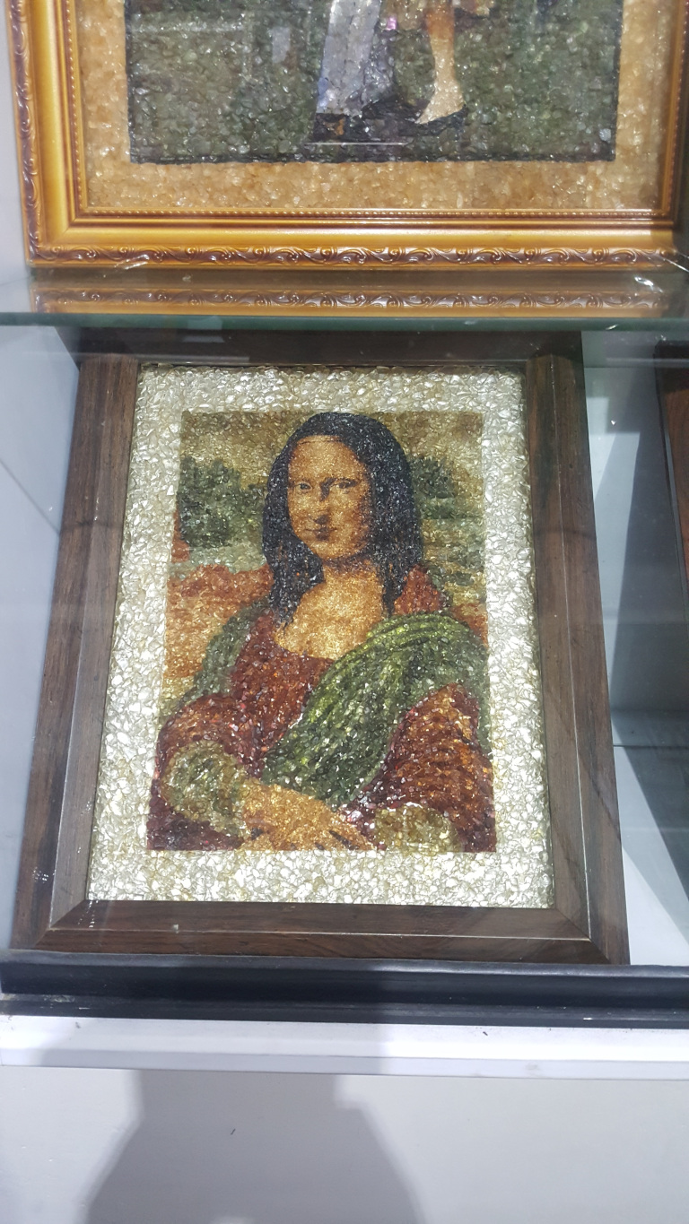 Rectangular frame holding a mosaic-style reproduction of the Mona Lisa. The Mona Lisa is depicted using what appears to be small, irregularly shaped pieces of amber or a similar translucent material, ranging in tones from deep orange-brown to golden-yellow and interspersed with smaller pieces of silvery-white material, creating a textured and shimmering effect. The background of the Mona Lisa mosaic is a muted mix of browns and greens, also created with the same textured material. The frame itself is a dark brown wood with a relatively simple, slightly bevelled profile. The setting is clearly indoors, likely a shop or gallery. The lighting is soft and even, avoiding harsh shadows and allowing the texture and colour variations of the mosaics to be clearly visible. The colour palette is warm, dominated by the earth tones of the frames and the amber-like material, contrasted by the silvery-white highlights within the mosaics. The white background provides a neutral backdrop, ensuring the artworks are the central focus.