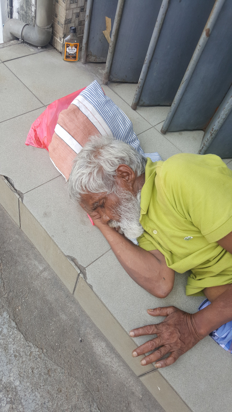 Elderly man with long, greying white hair and a full, white beard, lying asleep on a tiled walkway. He's resting on his side, his head supported by his left arm, which is bent beneath his cheek. His right arm is extended outwards, palm down, on the ground. He's dressed in a light lime green polo shirt, and a glimpse of blue and white patterned fabric is visible underneath the shirt, suggesting a pair of pants or a sarong. His skin is deeply tanned and shows signs of age and sun exposure. His expression is peaceful but his countenance suggests weariness or exhaustion. Near his head, a red plastic bag partially holds up a striped pillow providing some cushioning. A partially visible, clear, square-shaped bottle resembling a liquor bottle lies near the wall behind him.