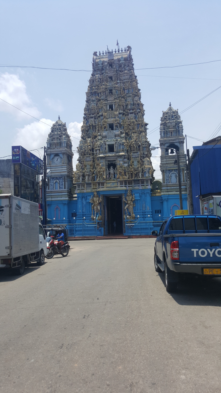 Large, ornate Hindu temple, predominantly light blue with intricate gold and beige detailing on its gopuram (tower). The gopuram is densely covered in carvings depicting numerous figures, likely deities and mythological scenes. Two smaller, similarly styled towers flank the main gopuram. In the foreground, a blue Toyota pickup truck is parked on the right side of a street facing away from the viewer. A motorbike is parked on the left, facing away. A light grey delivery truck is partially visible on the far left, also facing away. A few people are vaguely discernible within the temple entrance, but are too distant and small to provide detailed descriptions. The scene is set on a street, possibly in an urban or suburban area. Buildings are visible in the background, some of which have modern, commercial appearances. The sky is clear and light blue, indicating a sunny day. The lighting is bright and even, suggesting daytime photography under natural light.