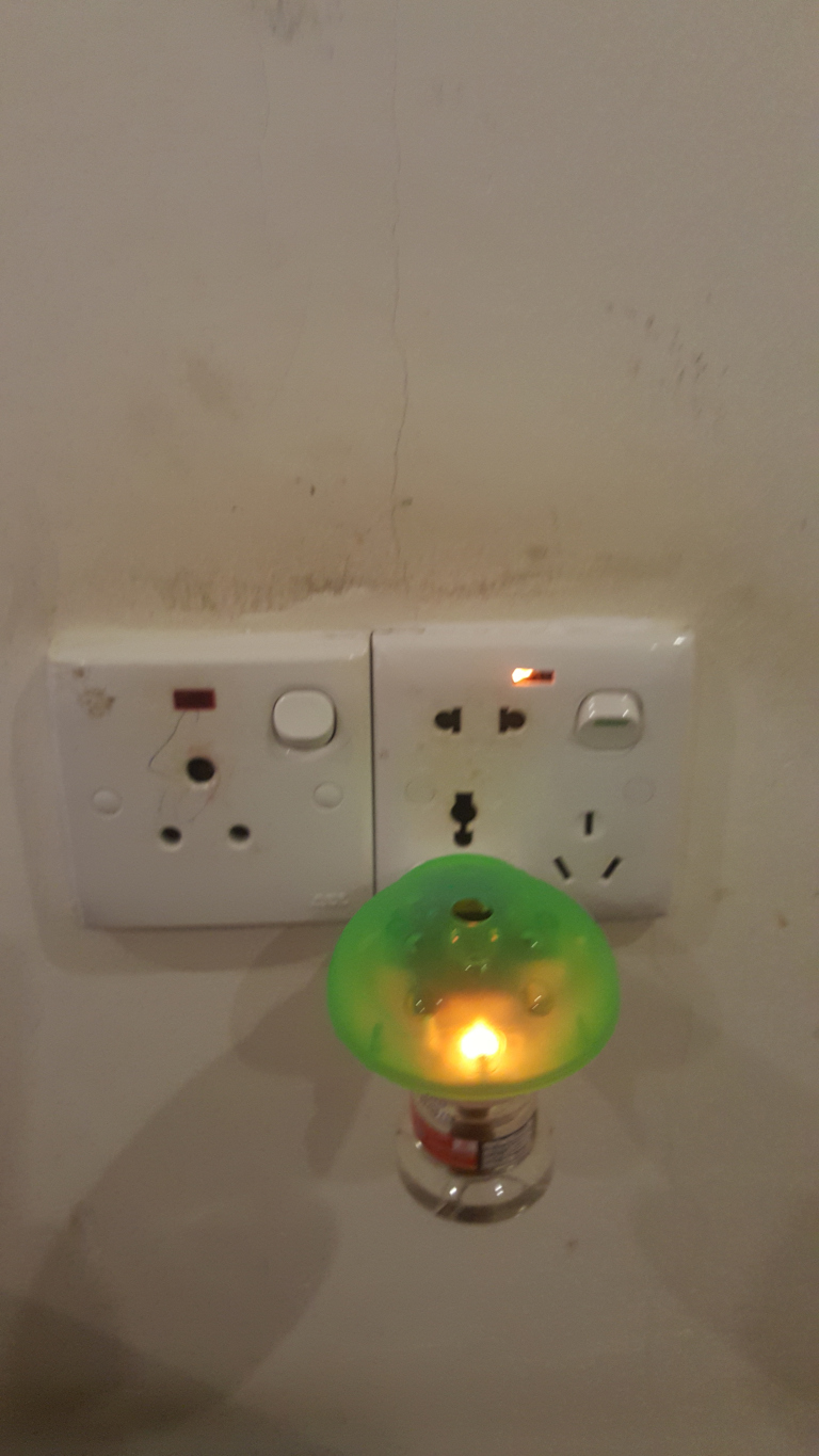 Nightlight with a lime-green plastic shade, resembling a stylised mushroom cap, emitting a warm, yellowish light from its base. The light is plugged into a double wall socket, which is a standard, off-white color and shows some signs of age and wear, including minor discolouration and what seems to be a slight crack or damage on one of the sockets. The switches on both sockets are off. There are no people present. The background is a light beige or off-white wall with noticeable imperfections—subtle discoloration, small blemishes, and a faint vertical crack running down its surface. The overall lighting is subdued, likely indoor ambient lighting, with the nightlight’s glow providing the brightest element of the scene. The colour palette is muted and warm, dominated by the off-white of the wall and the subtle yellowish glow of the nightlight against the lime green shade.