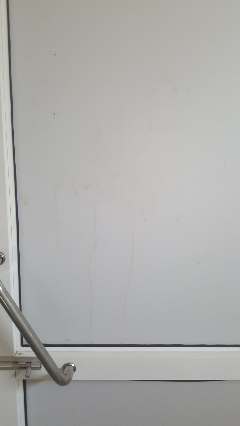 Light gray, possibly metal or composite, door or wall panel. The panel exhibits several vertical streaks of discoloration, possibly water stains or dirt, running from top to bottom in an uneven pattern. The stains are light brownish-gray, a slightly darker shade than the background. At the bottom of the panel, a horizontal white frame separates the panel from what appears to be the lower portion of the door or wall. A brushed stainless steel grab bar, angled slightly downwards, is affixed to the frame. A simple latch mechanism is visible on the door frame near the grab bar.