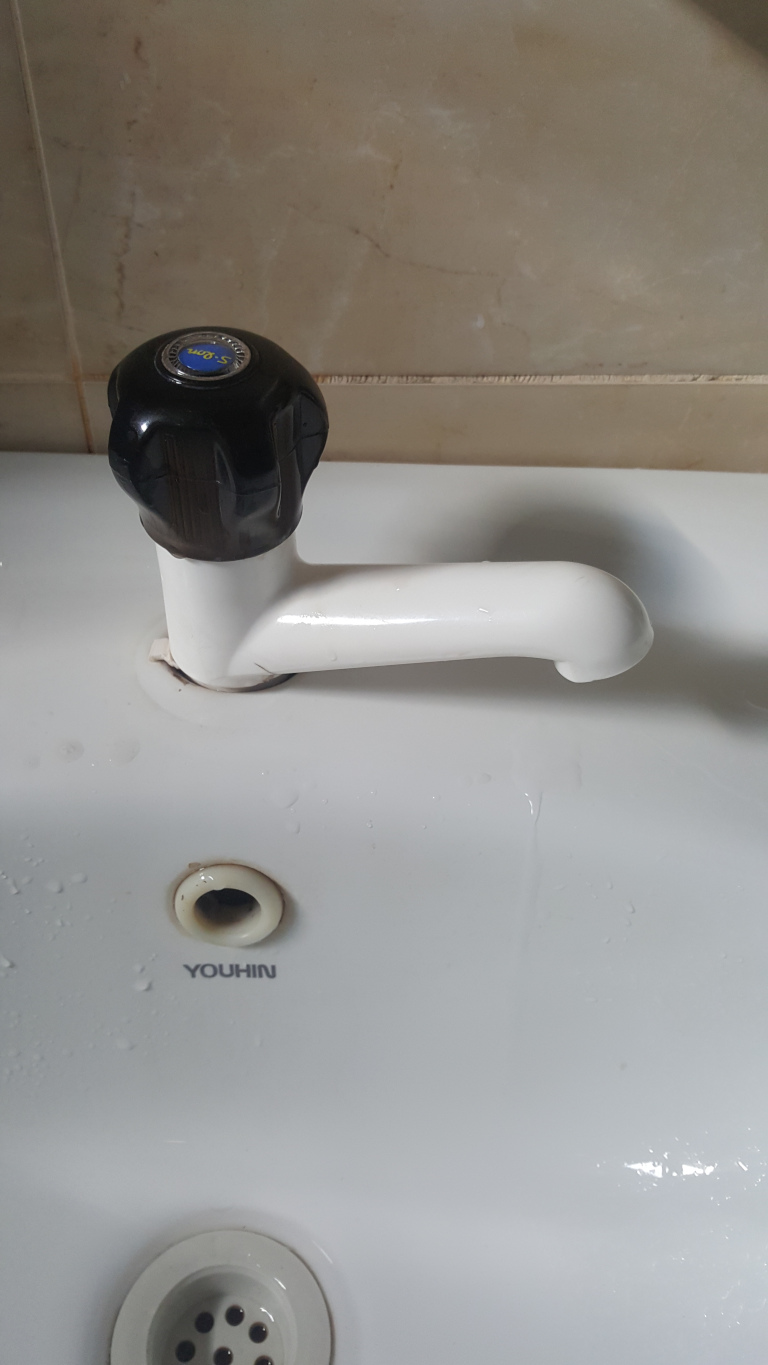 White bathroom sink with a single, slightly off-centre faucet. The faucet is predominantly white, with a dark brown, knurled, circular handle on top. The handle displays a small, partially visible blue circular logo. The faucet's spout extends horizontally, curving slightly downward. The sink shows minor water droplets and streaks on its surface, indicating recent use. Below the faucet is a small, round overflow drain, and further down is the main drain, visible as a circular metal grate with small holes. The word YOUHIN is printed in light gray on the sink's surface, below the overflow drain.