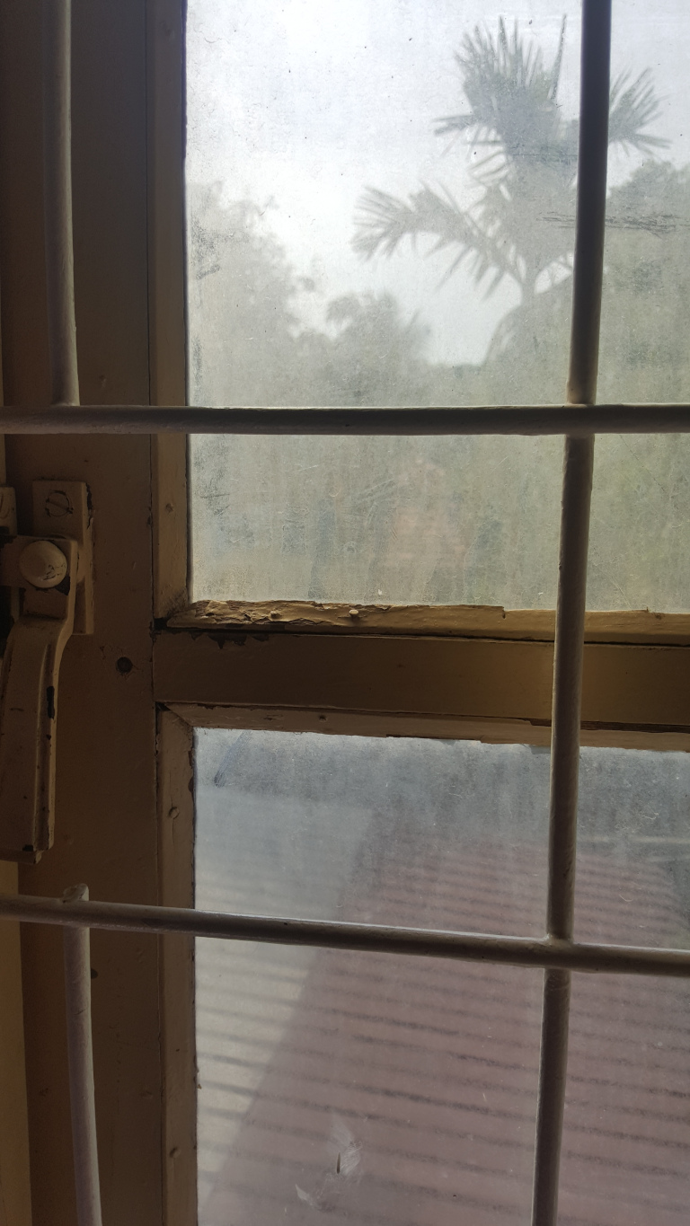 Dirty, multi-paned window with a security grille. The window panes are rectangular and divided by thin, off-white metal bars forming a grid. The window frame is a faded, yellowish-beige, showing significant wear, with chipped and peeling paint, particularly noticeable at the joints between the panes. A rusty, metallic latch or lock is visible on the left side of the frame, suggesting a mechanism to secure the window. No people are present.