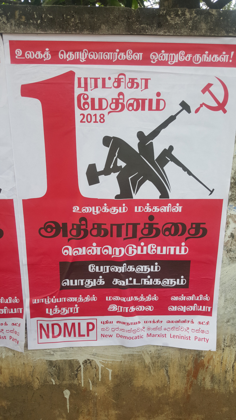 Vibrant red and white political poster, affixed to a weathered, textured wall. The poster is predominantly red, with white lettering and graphics. The poster's primary visual element is a stylised depiction of three figures, rendered in black silhouette, wielding hammers and a sickle, iconic symbols of communism. The figures are positioned dynamically, suggesting collaborative work or struggle. Above the figures, a large red 1 is prominently featured, likely symbolizing May Day (International Workers' Day). The hammer and sickle are also featured separately in red on the white upper section. Below the central image, there’s Tamil text, which appears to be the party's name, slogan, and other campaign-related information. At the bottom, NDMLP is displayed prominently in a rectangular box, accompanied by a translation indicating New Democratic Marxist Leninist Party. Additional Tamil text specifies locations for rallies or meetings.