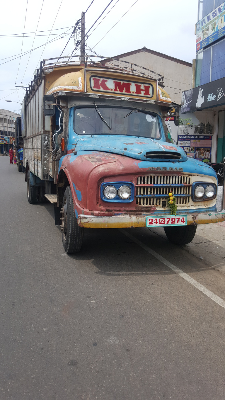 Vintage, heavily worn-down truck, prominently featuring the letters KMH on its yellow-cream cab. The truck's body is a patchwork of faded blue, red, and cream paint, showing significant signs of rust and age. Its headlights are round and slightly yellowed. The truck has a wooden cargo area. A small bouquet of yellow flowers adorns the grill. The license plate, 247974, is visible.Several people are visible in the far background, small and indistinct, suggesting a busy street scene. They are too far away to make out any details about their appearance or actions.