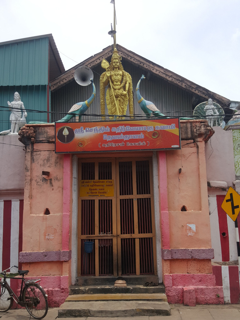 Hindu temple entrance, ornately decorated. Above the entrance, a golden statue of a Hindu deity, likely Lord Murugan (given the peacock imagery), stands majestically. The deity is adorned with intricate jewelry and holds what appears to be a spear or weapon. Two large, vibrant peacock statues flank the central figure. Smaller, white statues of what seem to be other deities or guardians are positioned on either side of the entrance at ground level. A large, rectangular sign bearing Tamil script is prominently placed directly above the temple door. A simple wooden door with vertical bars forms the main entrance to the temple. A bicycle is parked to the left of the temple structure, suggesting a street scene.