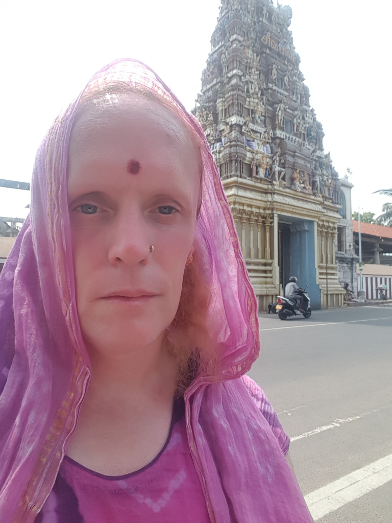 Leonie with light blonde hair partially visible beneath a draped, lilac-pink shawl or scarf. The shawl, which has subtle gold detailing along its edges, is loosely wrapped around her head and shoulders, partially obscuring their hair and framing their face. She has a prominent, round forehead with a small, dark red bindi centrally placed between her eyebrows. Her expression is serious, almost pensive, with a neutral, slightly downturned mouth. A small, light-coloured piercing is visible in her left nostril. She is wearing a purple tie-dye-style shirt underneath the shawl. She appears to be standing on a street, facing the camera directly. In the background, slightly out of focus, is a person on a scooter, moving away from the camera.