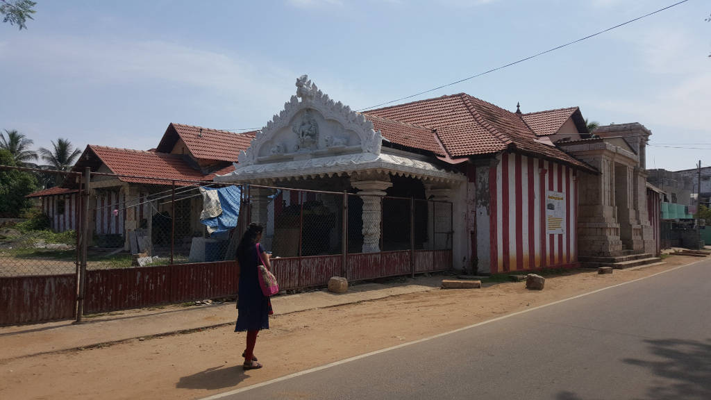 Partially dilapidated Hindu temple, characterised by a contrasting architectural style. The front section features a bright white, intricately carved gable and pillared portico, suggesting a more recent renovation or addition. This contrasts sharply with the adjacent, older sections of the building, which are clad in reddish-brown tiled roofs and painted with bold, vertical red and white stripes. The building shows signs of age and wear; some of the white paint is chipped or stained, and the red and white stripes are faded in places. A woman, seemingly young, stands facing away from the camera slightly to the left, in front of the temple’s rusty red metal fence. She is dressed in a dark blue, knee-length dress and crimson leggings, carrying a pinkish-purple handbag. She appears to be observing the temple.