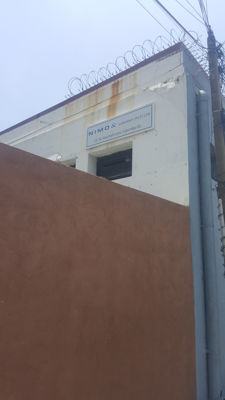 Building's facade, specifically a rectangular sign affixed to its upper portion. The sign, a crisp, almost clinical blue and white, reads NIMO & COMPANY (PVT) LTD in a sans-serif typeface, underneath which is the address: 55/16, Vauxhall Lane, Colombo 02. The building itself is predominantly white, showing signs of age and weathering, particularly a rust-colored discoloration along its upper edge beneath a rusty barbed wire fence. A large, deep brown stucco wall extends from the main building, forming a significant part of the composition's left side. A dark grey pipe or conduit runs vertically along the right edge of the white building section. The sky is a clear, pale blue, almost washed out, suggesting midday sunlight.