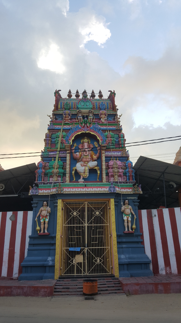Vibrant, intricately detailed Hindu temple entrance. Dominating the entrance is a large, colourful statue of Ganesha, the elephant-headed god, seated on a throne. He is depicted in a traditional pose, holding his attributes. The statue is brightly painted with reds, greens, blues, and golds. Above Ganesha, a gopuram—the characteristic tower of a South Indian temple—rises. It is multi-tiered and richly decorated with smaller statues of deities and mythical figures, all rendered in a similar bright colour palette. Intricate carvings and decorative elements abound.