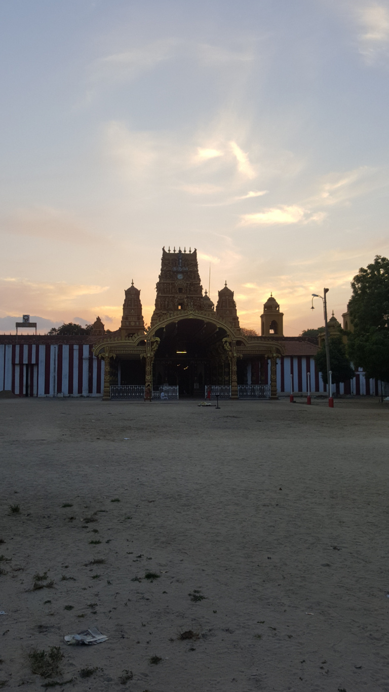 Large, ornate Hindu temple, its gopuram (tower) dominating the scene. The gopuram is golden-hued, richly decorated with intricate carvings and details, appearing almost jewel-toned in the twilight light. Two smaller, flanking gopurams are visible on either side of the main structure. The temple's entrance is a large, shadowed archway. A few indistinct figures can be seen near the entrance, but they are too small to discern any details. The temple sits behind a low wall with alternating red and white vertical stripes. The foreground is a mostly empty, flat, sandy or dusty area. A single piece of discarded paper is visible near the bottom left. The scene is set outdoors, in what appears to be a temple courtyard or open space. The background features a sparse, low-lying horizon line with a few scattered trees visible to the right. The sky is a soft pastel blend of pale blues, pinks, and oranges characteristic of twilight or sunrise. The clouds are thin and wispy, with areas of lighter and darker tones.