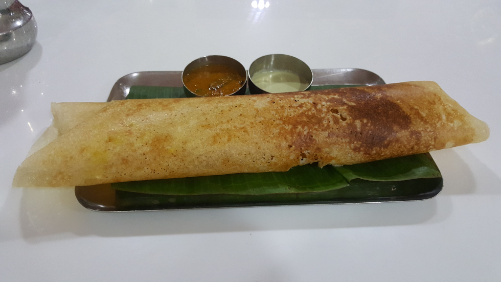 Masala dosa, a South Indian crepe, presented elegantly on a rectangular stainless steel tray. The dosa is light golden-brown, slightly browned in places, suggesting it's been cooked on a hot griddle. It's rolled up loosely, revealing a soft, almost creamy interior. It rests on a vibrant green banana leaf, partially visible underneath, adding a touch of natural texture and color contrast. Two small, stainless steel bowls are positioned in the upper middle of the tray. One contains a bright yellow-orange sambar (a lentil-based vegetable stew), while the other holds a light green chutney (a condiment). The position and slight overlap of these elements guide the viewer’s eye across the composition.