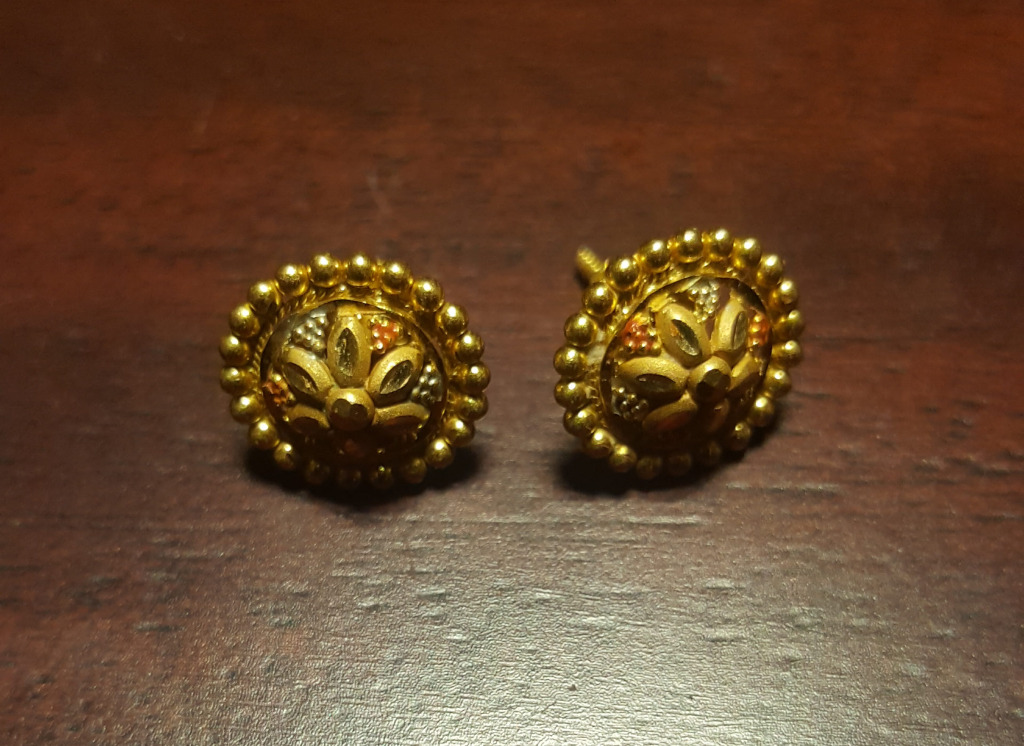 Earrings are the focal point, positioned slightly off-centre, with one earring subtly ahead of the other. They are small, round, and gold, featuring a central floral design intricately detailed with what appears to be multi-colored gold (perhaps different karats or colored gold inlay). The floral motif is composed of several stylized petals radiating from a central point, accented with tiny specks of color, possibly red, white, or silver. The central circle is encircled by a row of small, evenly spaced golden beads, creating a textured halo effect. Each earring has a small, almost invisible post visible at the top.