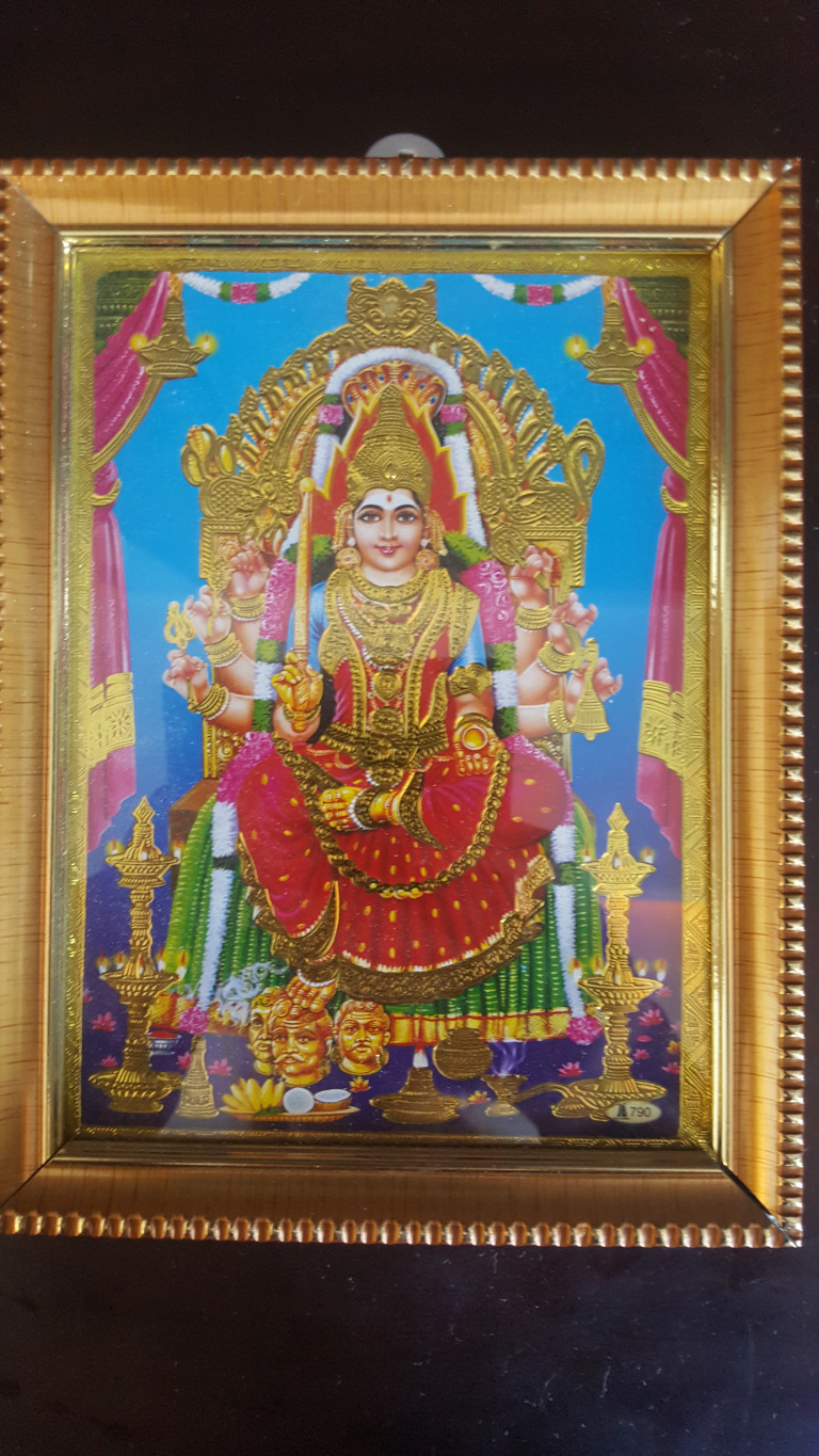 Richly adorned goddess seated majestically on a golden throne. She is the focal point, occupying most of the frame. Her attire is extravagant: a vibrant red and gold sari embellished with intricate patterns and sparkling details, suggesting opulence and divinity. Her complexion is light, and her expression is serene yet regal. She wears heavy gold jewelry, including necklaces, bangles, earrings, and a crown, all meticulously detailed. Her multiple arms (characteristic of many Hindu goddesses) are gracefully posed, holding various symbolic objects – possibly a weapon, a flower, or a blessing gesture.  At the bottom of the image, three severed male heads are depicted, a common motif in some depictions of the goddess, symbolising the triumph of good over evil.