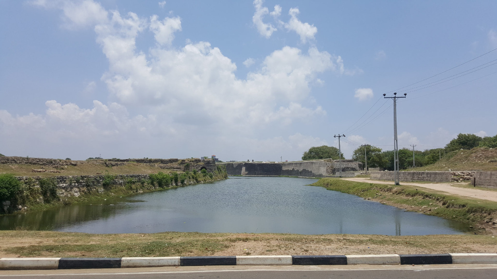 Tranquil moat, a long, narrow body of still, dark-greenish water, reflecting the sky and the surrounding structures.  The moat is bordered by low, grassy banks and runs parallel to a section of ancient, weathered stone walls, which form a low, fortress-like structure. These walls, a muted gray-brown, show signs of age and wear, with patches of vegetation sprouting from cracks and crevices.  Beyond the wall, a flat, grassy expanse extends to the horizon. Power lines stretch across the right side of the image, leading the eye towards a slightly elevated area with sparse, low-lying trees. The foreground features a simple, black-and-white painted curb along a paved road, separating the viewer from the moat. No people or animals are visible.