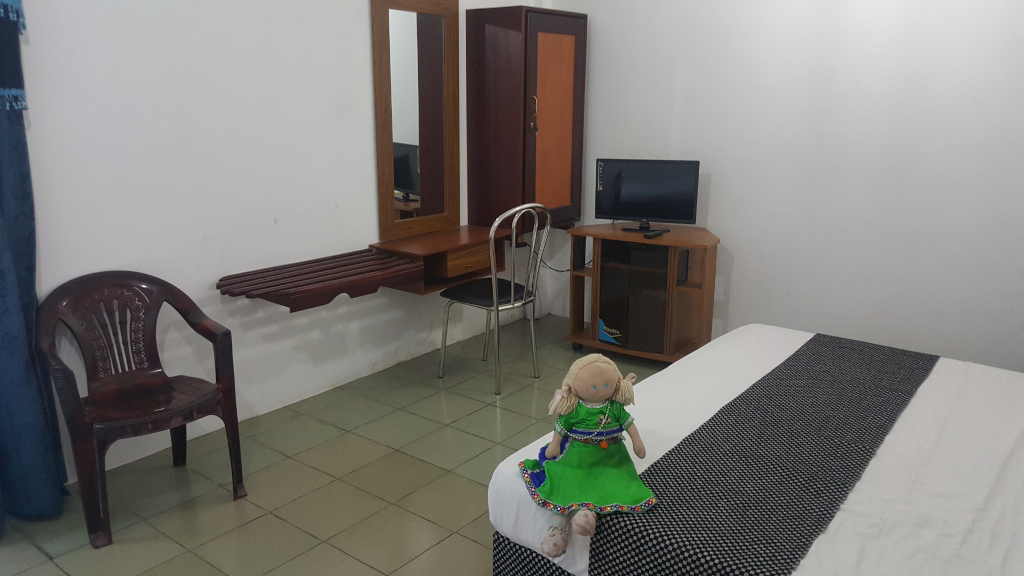 Seated centrally in the foreground on a bed, slightly off-center to the left. The doll is fair-skinned with blonde yarn hair, wearing a vibrant lime green dress adorned with intricate, dark-colored embroidery that resembles traditional South Asian or folk styling.  She wears a simple necklace. The doll is posed in a slightly relaxed manner, arms slightly out to the sides. A white bed with a black and white checkered runner dominates the bottom right of the image. The bedding is crisp and appears relatively new. A dark brown plastic chair with ornate detailing sits to the left, near the vanity area, appearing slightly worn or used. The scene is a modestly furnished bedroom. The walls are plain white, emphasising the furniture. The floor is light-coloured tile, appearing clean but simple.