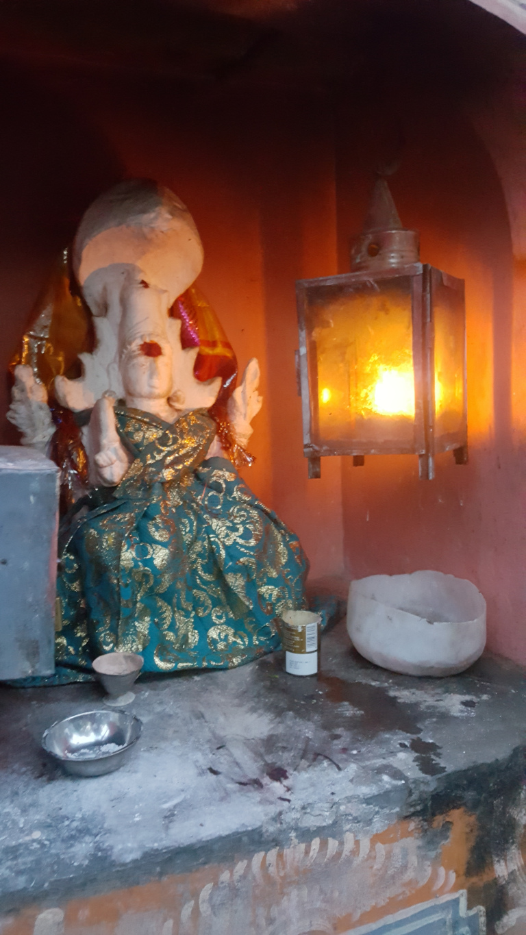 Small, intricately dressed statue of what appears to be a Hindu deity, possibly a form of Ganesha. The statue is predominantly white or light beige, with a rounded head and what seems like elephantine features, though partially obscured. It’s adorned in a vibrant teal and gold patterned garment, draped in flowing folds. The dress is richly decorated with gold accents, giving a sense of opulence. The statue's hands are posed in a traditional devotional gesture. To the right, a simple, hanging lantern made of glass and metal casts a warm, orange glow. Inside, a small flame burns brightly, illuminating the scene.  Near the base of the statue, there are small bowls, one metallic and possibly containing ash or some other offering, and another white bowl which could be used for offerings. A small, cylindrical container, possibly a small tin or candle, sits near the lantern.