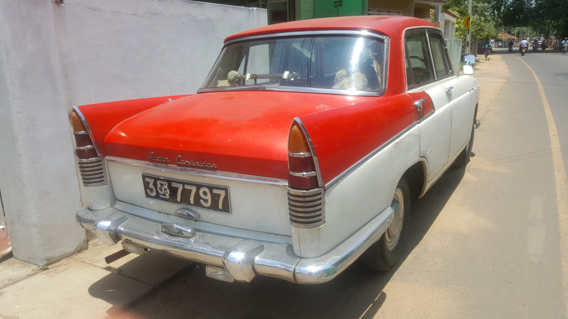 Vintage Austin Cambridge car, predominantly white with a vibrant red roof. The car is parked on the side of a road, angled slightly away from the viewer, showing its rear three-quarter view. The car appears slightly dusty or weathered, suggesting age and use. Its chrome detailing, particularly the bumpers and tail lights, are slightly tarnished but still reflective, catching the sunlight. The license plate, 38 7797, is clearly visible. Inside the car, two light-coloured, possibly stuffed animal, objects are partially visible in the rear seat. There's no human presence directly interacting with the car, although distant figures are visible in the background.