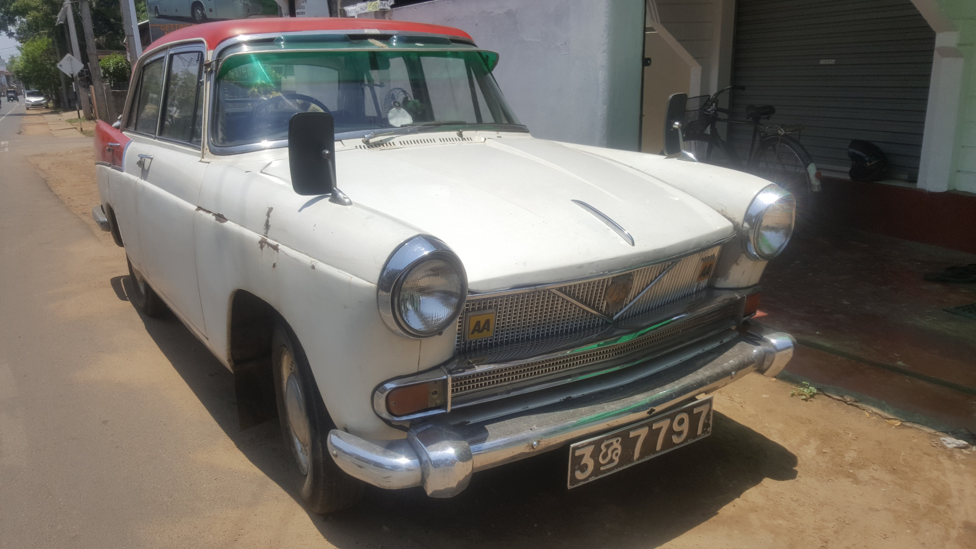 Vintage, classic car, seemingly a 1960s model, predominantly white with a faded red roof. The car shows signs of age and wear; the paint is chipped and faded in places, and there’s a noticeable patina of age. It's parked on the side of a street, angled slightly away from the viewer. The car has chrome bumpers, grill, and window trim that sparkle in the sunlight. A license plate, 38 7797, is prominently displayed on the front bumper. The car's side mirrors are visible, and a small, yellow AA sticker (possibly an auto association logo) is on the grill. The scene is set on a sun-drenched street in what appears to be a residential area in a tropical or subtropical climate.  The street is paved but shows some discoloration, indicating dust or possibly sand. The background includes a simple, light-colored building with a roller door, partially visible on the right. A bicycle is leaning against the building. The buildings in the far distance are hazy and indistinct. The lighting suggests it’s likely midday, with bright, harsh sunlight casting distinct shadows.
