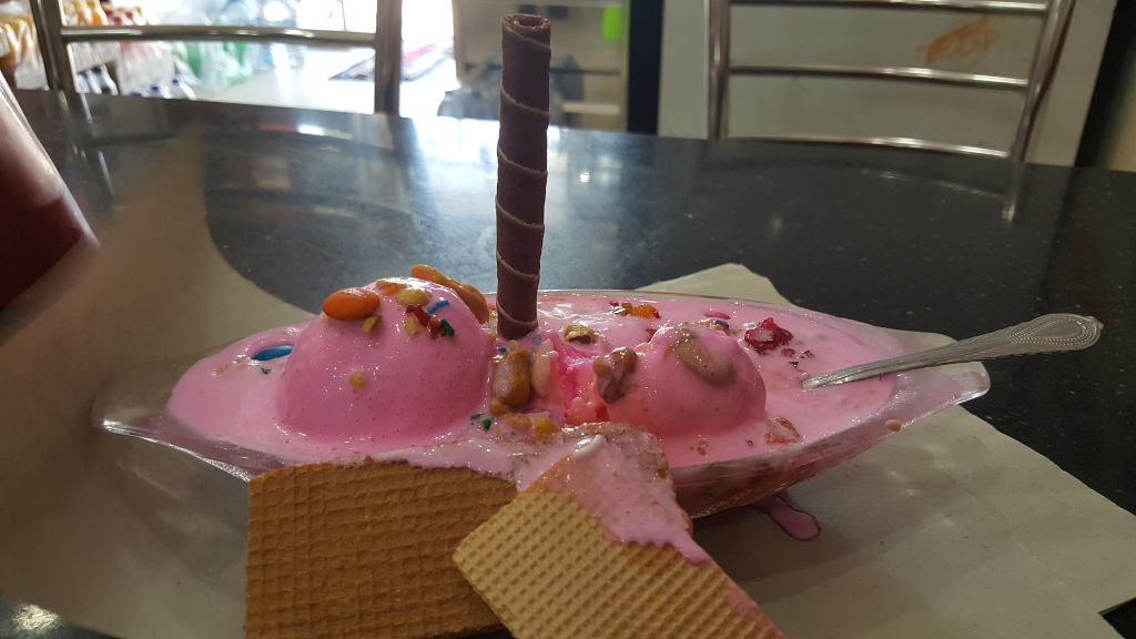 Vibrant pink ice cream sundae, overflowing from a clear, glass boat-shaped dish. Two generous scoops of pink ice cream are nestled within a pool of pink sauce or melted ice cream. The ice cream is generously adorned with colourful sprinkles, chopped nuts (cashews appear prominent), and what look like small, colourful candy pieces. A tall, dark chocolate wafer stick stands upright in the centre of the dessert, adding a vertical element to the composition. A portion of a light brown waffle or biscuit is visible on the bottom left, with another broken piece partially submerged in the melted ice cream on the right, suggesting the ice cream has been partially eaten. A silver spoon rests alongside the dish, appearing to have been recently used.